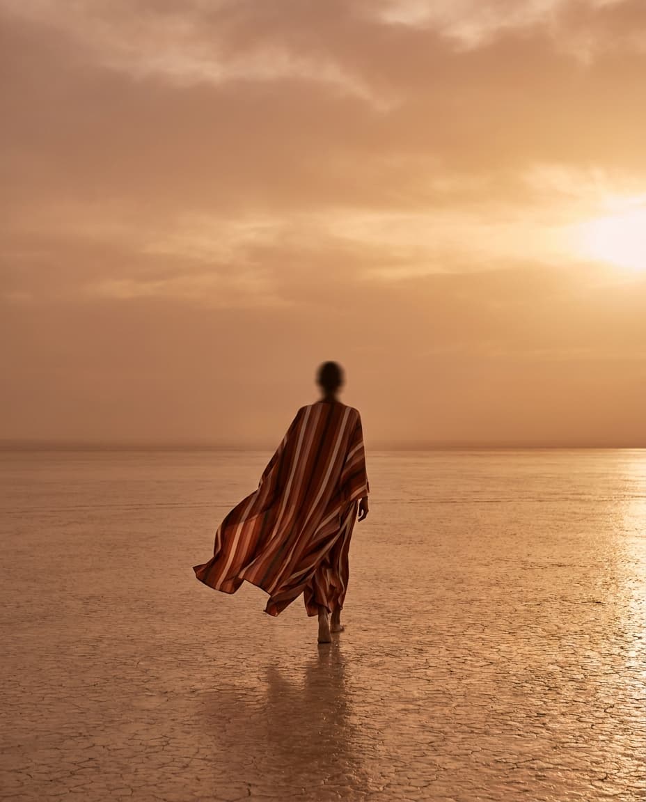 Anonymous African figure in a striped wrap walking across a cracked-earth salt flat at golden hour